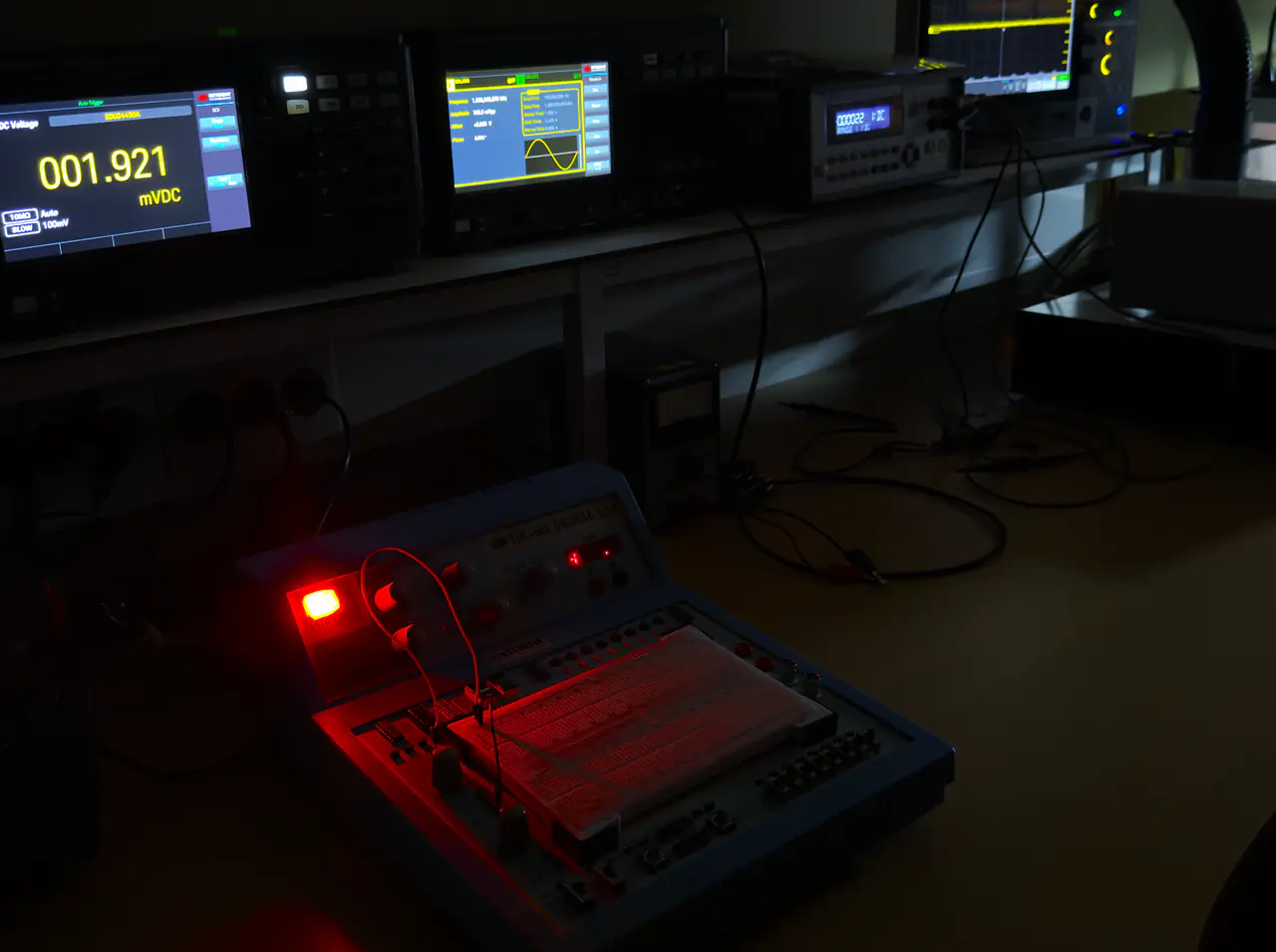 En arbeidsbenk i mørket: et multimeter viser 001.921 mVDC, og et koblingsbrett for digitalteknikk lyser rødt i forgrunnen.