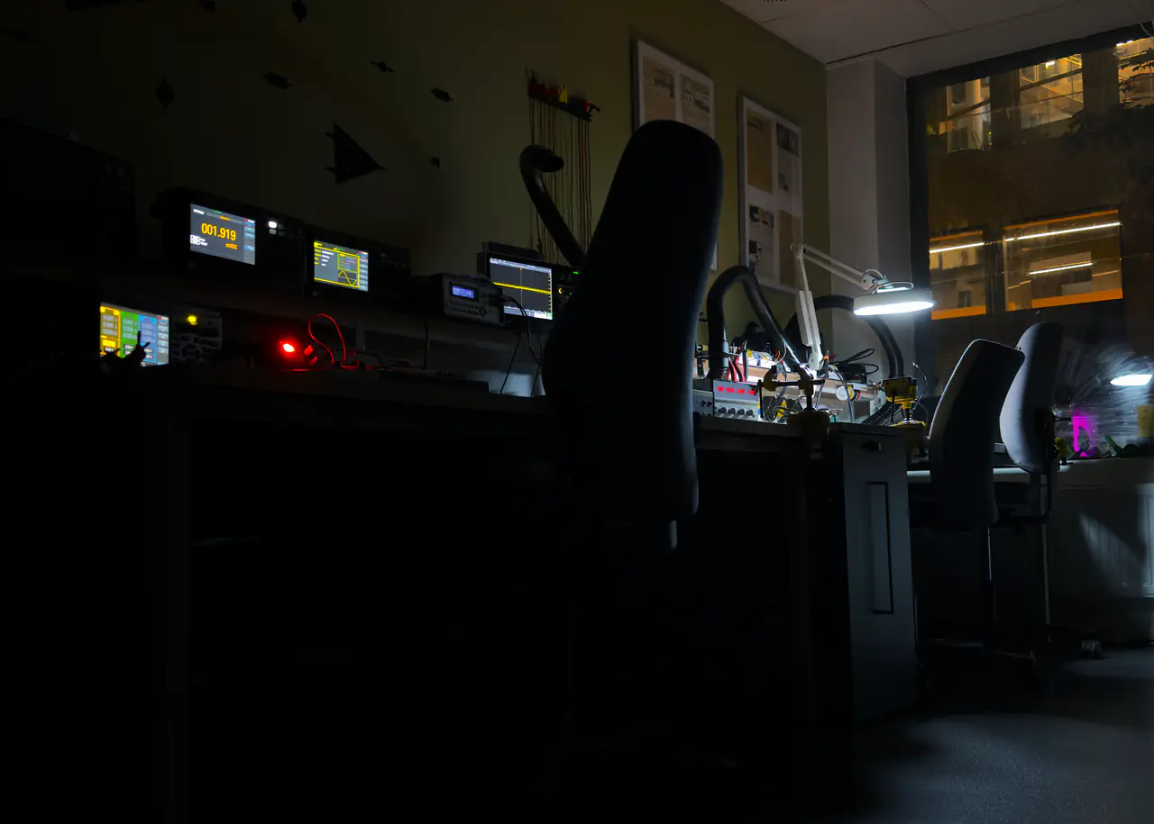 Verkstedet om kvelden: skjermene på multimetre, signalgeneratorer og oscilloskop lyser langs arbeidsbenken, og en arbeidslampe står tent ved vinduet.
